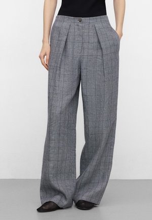 Vrouw met een grijze geruite wijde broek met hoge taille, voorplooien, zakken, knoopsluiting, een zwart topje en zwarte puntige schoenen.