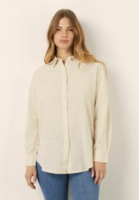Camisa de color crema con cuello puntiagudo, mangas largas y un corte relajado. Fabricada con un tejido ligero y con una textura sutil.