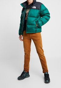 Groene pufferjack met zwarte accenten, met ritssluitingen, gedragen over een zwart graphic t-shirt. Gecombineerd met bruine broek en zwarte sneakers.