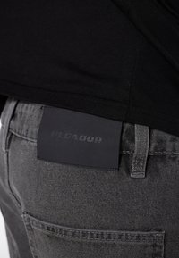 Gray denim jeans with a textured fabric, featuring a black leather tag labeled "PEGADOR" on the back. Simple design and neutral color.
