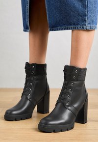 Bottes noires à la cheville en cuir synthétique, dotées d'un talon bloc épais, d'un laçage frontal et d'une tige texturée avec des accents brillants.