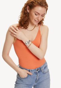 Rippend orange Bodysuit mit dünnen Trägern, kombiniert mit hellblauen Jeans. Eine silberne Uhr und eine goldfarbene Kette setzen Akzentdetails.