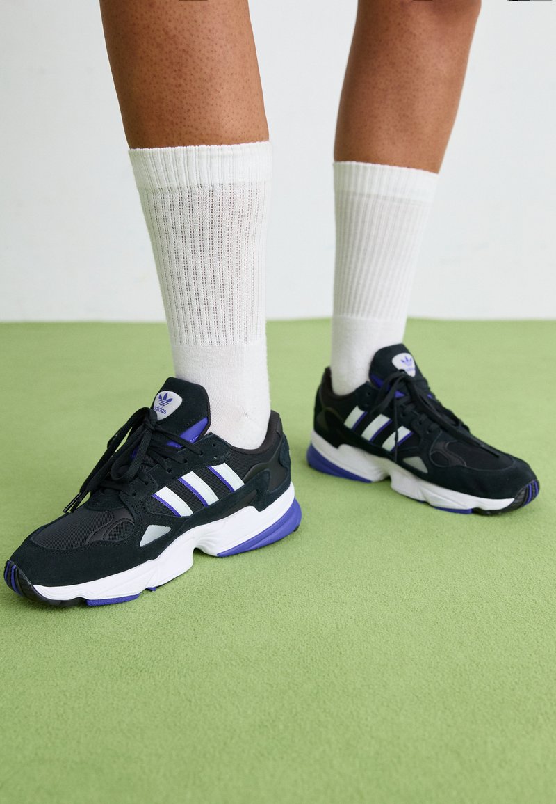Czarne buty sportowe z białymi i fioletowymi akcentami, wykonane z połączenia siateczki i zamszu, na zielonym tle.