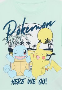 La camiseta turquesa presenta una impresión gráfica de una tortuga azul sonriente y un ratón amarillo. El texto dice "Pokémon" sobre un atardecer y palmeras.