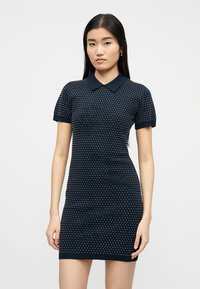 Γυναίκα που φοράει μπλε ναυτικό, κοντομάνικο, πλεκτό φόρεμα με γιακά και μικρές λευκές πουά, στέκεται μπροστά σε απλό ανοιχτό φόντο.