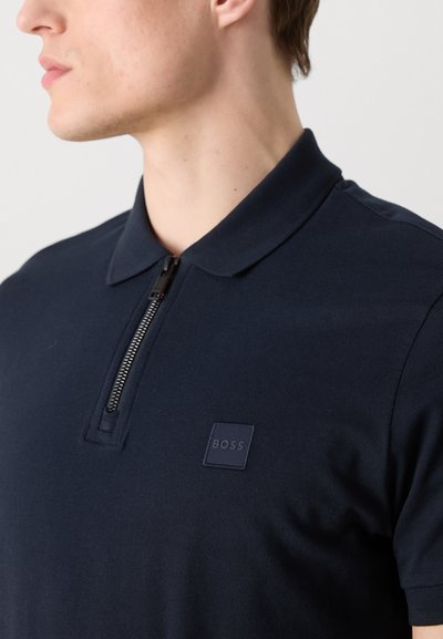 Polo shirt navy avec un col zippé, présentant un tissu texturé et un petit patch logo carré navy sur la poitrine gauche.