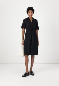 Abito nero button-up con un davanti annodato, maniche corte e orlo medio. Abbinato a una borsa bianca con un motivo trapuntato e mocassini neri.