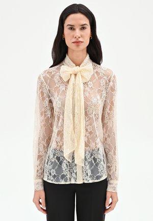 Femme portant une blouse en dentelle crème transparente à manches longues avec un grand nœud papillon au col, associée à un pantalon noir, debout devant un fond uni.