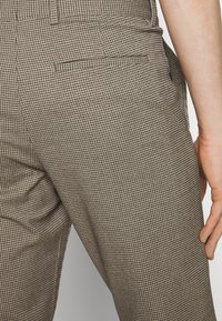 Brązowe, w kratę spodnie o dopasowanym kroju, z teksturowanej tkaniny i detalem w postaci tylnej kieszeni; wzór kolorystyczny składa się z małych, naprzemiennie ułożonych kwadratów.