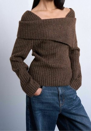 Femme portant un pull côtelé marron à épaules dénudées, associé à un jean bleu taille haute, mains dans les poches, sur fond uni.