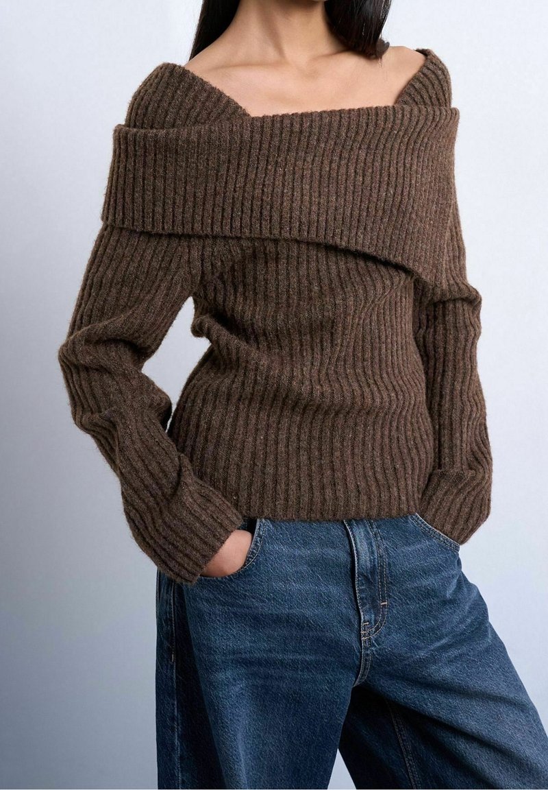 Femme portant un pull côtelé marron à épaules dénudées, associé à un jean bleu taille haute, mains dans les poches, sur fond uni.