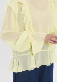 Gelbe Chiffon-Bluse mit Rüschendetails, langen Ärmeln und geschichtetem Saum. Leichte, durchsichtige Textur und figurbetonte Bündchen unterstreichen das Design.