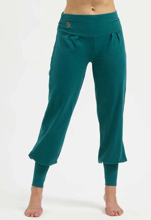 Frau trägt türkisfarbene geschichtete, weit geschnittene, kurze Hosen mit enganliegenden Leggings darunter und steht barfuß auf weißem Hintergrund.
