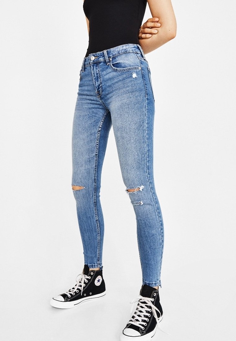 Tunna blå denimjeans med slitna knäskärningar, en figursydd siluett och medelhögt midjan. Kombineras med svarta högstövlar.