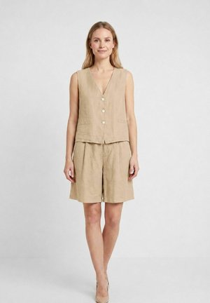 Kvinde står iført beige ærmeløs vest med knapper og matchende knælange shorts med neutrale hæle mod en ensfarvet baggrund.