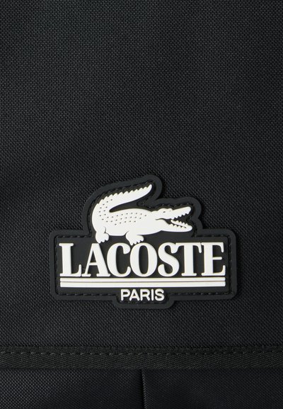Черен плат с бял нашивка с лого на Lacoste Paris, с крокодил над името на марката, пришит върху повърхността.