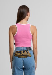 Sieviete ar gariem brūniem matiem, kas valkā rozā īso topu un zilos džinsus ar lielu dzeltenu "Von Dutch Originals" uzkali mugurpusē.