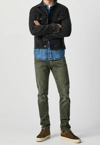 Black denim jacket over a blue chambray shirt, paired with green trousers and brown sneakers. Simple design with classic fit.