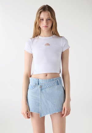 Giovane donna che indossa un top bianco corto con un piccolo ritaglio e una minigonna in denim azzurro asimmetrica, in piedi davanti a uno sfondo bianco.