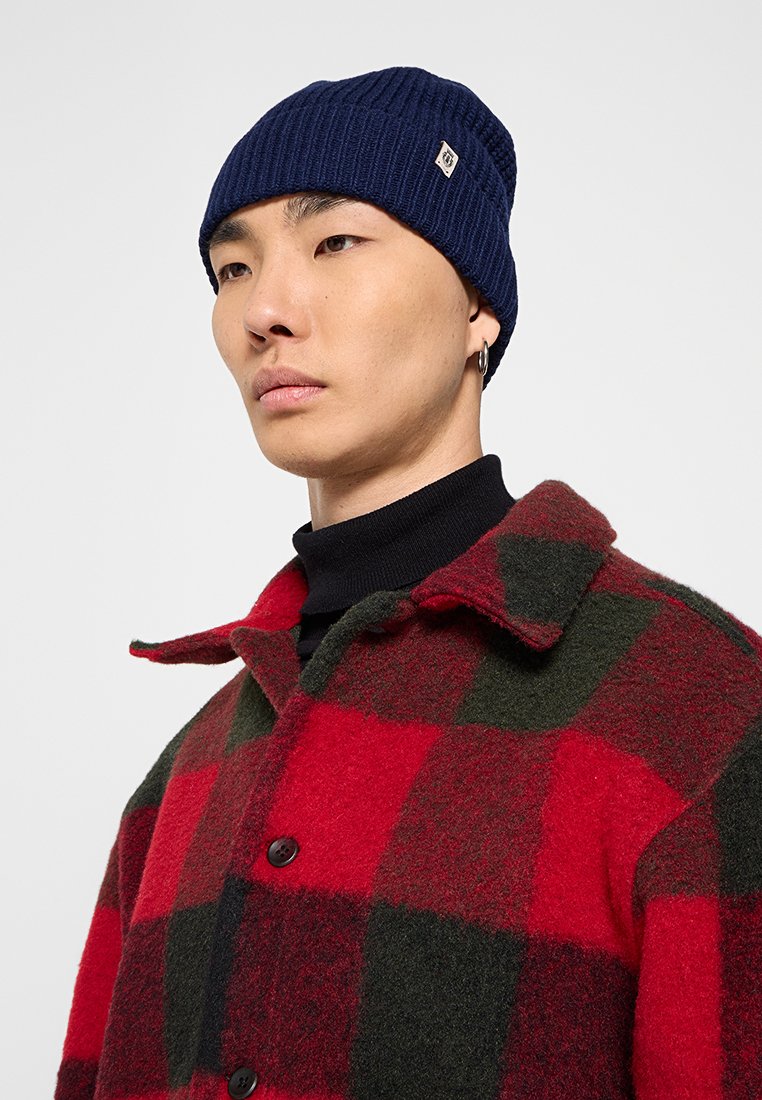 Mężczyzna noszący granatową dzianinową czapkę, srebrną kolczyk w kształcie kółka, czarny golf oraz czerwono-ciemnozielony wełniany płaszcz w kratkę z kołnierzem i guzikami.