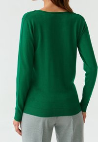 Maglione verde lavorato a maglia con scollo rotondo e maniche lunghe. Presenta orlo e polsini a coste, mostrato da dietro su uno sfondo neutro.