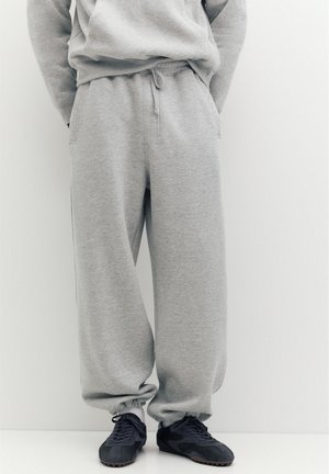 Personne portant un pantalon de survêtement gris ample et des baskets noires, debout devant un fond clair uni.