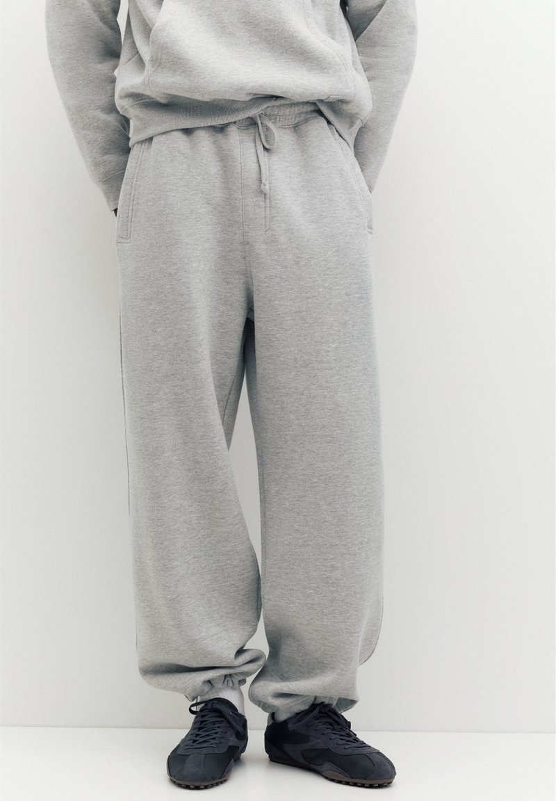 Personne portant un pantalon de survêtement gris ample et des baskets noires, debout devant un fond clair uni.