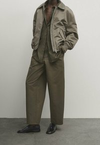 Olijfgroene bomberjack met een rits, gecombineerd met losse bijpassende broeken, gedragen over een donker shirt. Zwarte leren schoenen maken de look af.