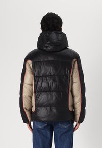 Casaco puffer com painéis pretos e bege, com costuras vermelhas em destaque. O casaco tem capuz e uma textura acolchoada.