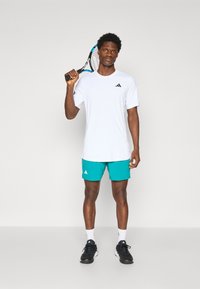Witte sport t-shirt met zwart logo, gecombineerd met turquoise shorts met witte accenten, zwarte sportschoenen en een tennisracket.