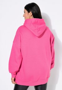 Kobieta z długimi ciemnymi włosami, nosząca duże kolczyki w kształcie kół i jasnoróżowy, oversize'owy hoodie, pokazana od tyłu na białym tle.