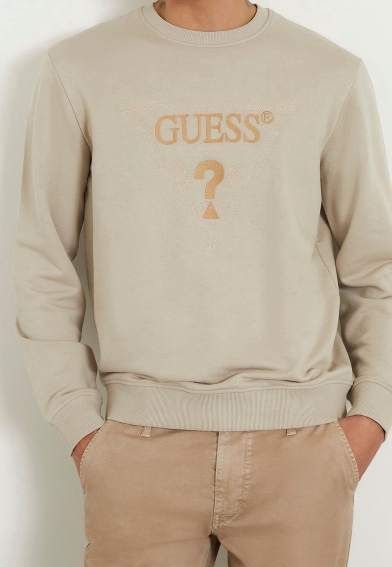 Sudadera de algodón beige con puños y dobladillo acanalados. Presenta un logotipo tonal "GUESS" y un diseño triangular con un signo de interrogación en el centro.