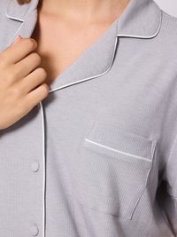 Grijze geribde pyjama top met witte piping langs de kraag en de zak. Heeft een knoopsluiting en een linkerborstzak. Zachte stoftextuur.