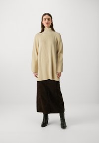 Beige, ribbet genser med høy hals og lange ermer, kombinert med en mørkebrun, kne-lang skjørt og svarte hælsko.