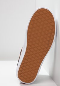 Sneaker-Sohle aus braunem, strukturiertem Gummimaterial mit Diamantmuster und weißen Seitenwänden. Vor hellem Hintergrund positioniert.