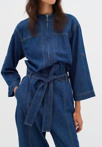 Dunkelblauer Denim-Overall mit hohem Kragen, Reißverschluss, langen Ärmeln und gebundenem Gürtel, mit sichtbaren Nähten und seitlichen Taschen.