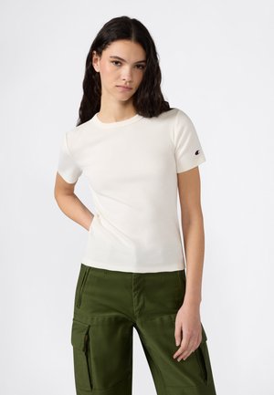 Giovane donna con capelli scuri ondulati che indossa una maglietta bianca a maniche corte e pantaloni cargo verde oliva, in piedi con una mano dietro la schiena.