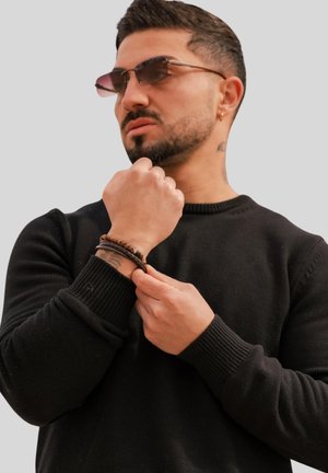 Black textured sweater, dark-toned bracelets on wrist, and sunglasses with gradient lenses. Background is neutral gray.