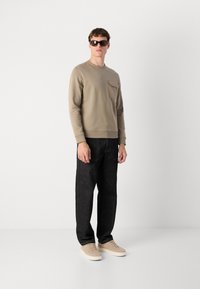 Sweatshirt castanho claro com um bolso no peito, combinado com jeans escuros e ténis bege. O modelo usa óculos de sol escuros e posa contra um fundo simples.