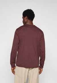 Camisa de manga longa em tecido bordeaux profundo. Apresenta um decote redondo e um corte relaxado, com textura suave e costuras ajustadas.