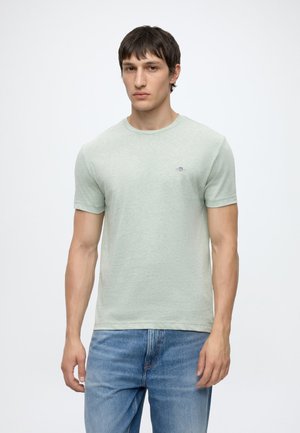 Jeune homme aux cheveux courts foncés, portant un t-shirt vert clair à manches courtes et un jean bleu, se tenant devant un fond blanc uni.