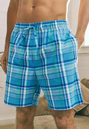 Blaue und grüne karierte Herrenshorts mit Kordelzugbund, getragen von einer oberkörperfreien Person, die drinnen steht.