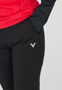 Sorte træningsbukser med en glat tekstur og tætsiddende design. Har et lille gråt logo på lårdetaljen og er parret med en rød top.