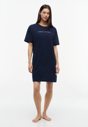 Kobieta stoi na boso, nosząc granatową sukienkę typu t-shirt Tommy Hilfiger z krótkimi rękawami na jednolitym białym tle.