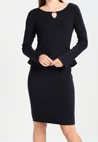 Robe tricotée bleu marine à manches longues, avec un ourlet à basque et un détail d'encolure en forme de goutte. Texture lisse, silhouette ajustée.