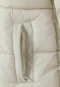 Heled beige puffer jakk, millel on ligipääsetav lähedalt, lukuga tasku valge tõmbluku pull tabiga, tikandite tekstuur ja sile kangas.