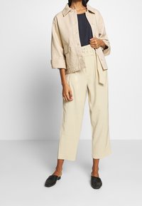 Beige kortjacka med stora knappar över en marinblå topp, kombinerad med högmidjade beige byxor och svarta flätade mules. Mjuka texturer.