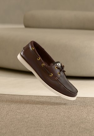 Chaussure bateau en cuir marron foncé avec semelle blanche et détails de lacets, posée sur un tapis beige devant un canapé beige.