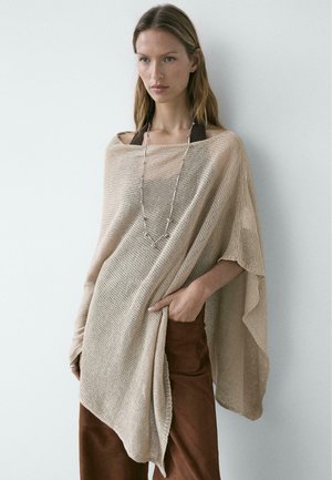 Donna che indossa un poncho lavorato a maglia beige sopra una maglietta scura e pantaloni marroni, accessoriata con una lunga collana d'argento, in piedi contro un muro bianco.