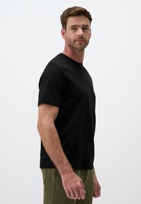 Czarna bawełniana koszulka z krótkim rękawem, okrągłym dekoltem i luźnym krojem, zestawiona z oliwkowymi spodniami, pokazana z boku.